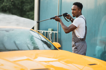 Manual car wash. Handsome African young man washing his luxury yellow vehicle with high pressure...