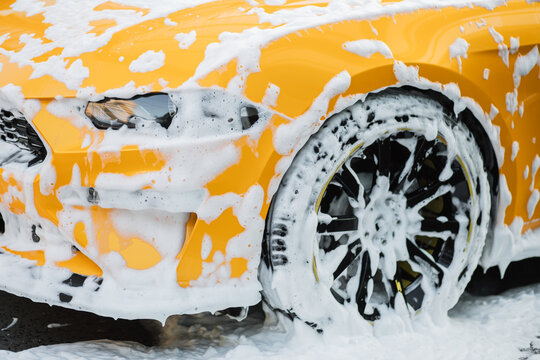 Cropped image of wheel of luxury yellow car in outdoors self service car wash, covered with cleaning soap foam.