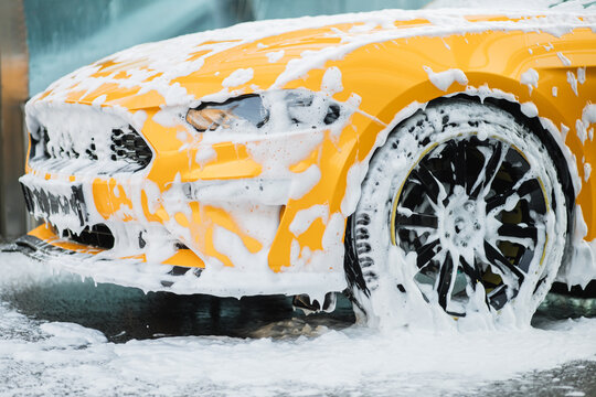 Cropped image of wheel of luxury yellow car in outdoors self-service car wash, covered with cleaning soap foam.