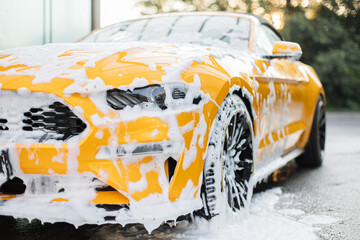 Cropped image of wheel of luxury yellow car in outdoors self-service car wash, covered with cleaning soap foam. - obrazy, fototapety, plakaty