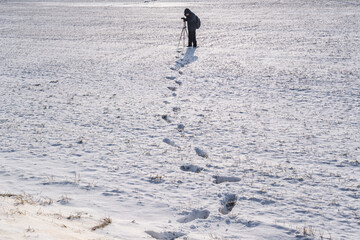 Fototapeta na wymiar photographer with a tripod in snowy field takes pictures of winter landscape, footprints in snow