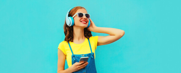 Portrait of happy smiling young woman in headphones listening to music with phone on blue background