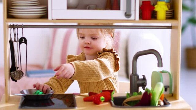 cute little baby girl plays on a toy kitchen, pretending to make lunch on a stove in the morning