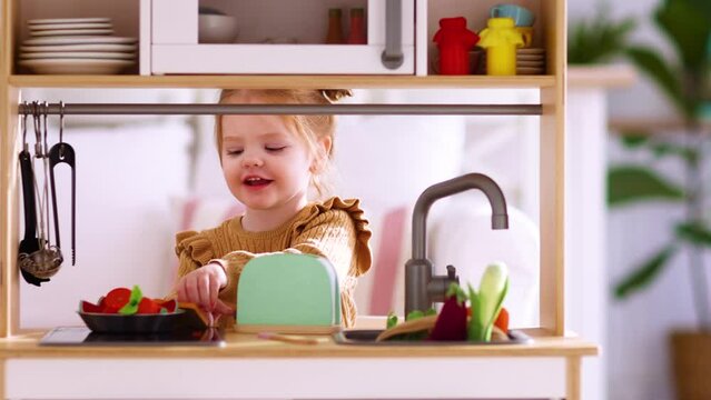 cute little baby girl plays on toy kitchen, pretending to make tasty toasts at sunny day