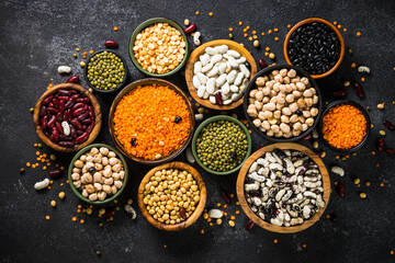 Legumes, lentils, chikpea and beans assortment in different bowls on black stone table. Top view with copy space. Vegan food, protein source.