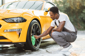 Car wash service outdoors. Car wash self-service concept. Handsome African guy wiping wheel of...