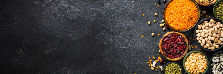 Legumes, lentils, chikpea and beans assortment in different bowls on black stone table. Long banner format.