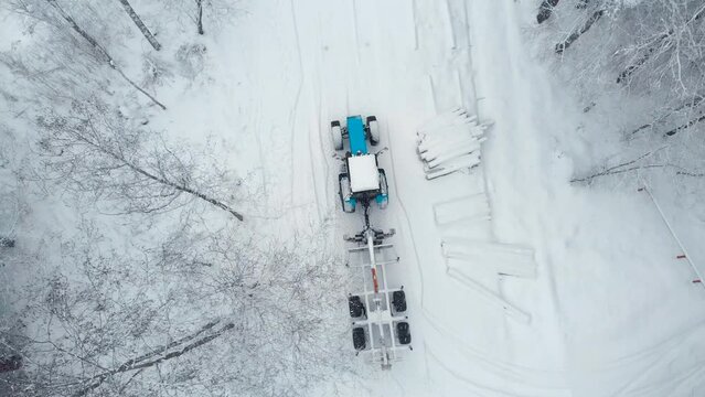Aerial view a tractor with a trailer for loading logs rides on a snow-covered road in the forest. Logs in the snow along the road.  Drone shooting 