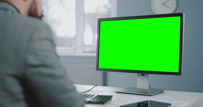 Back view of caucasian man in suit sitting at office desk and working on pc with chroma key monitor. Concept of people, business and technology.