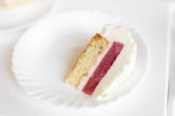 A white-pink piece of cake lies on a white plate on a light background. Delicious pastries