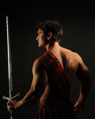 The sexy highlander is posing indoor wearing a medieval costume and holding a sword. 