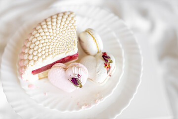 Obraz na płótnie Canvas A white-pink piece of cake lies on a white plate on a light background. A pink heart-shaped cake lies on a white plate on a light background. Light macaroons on a white canvas. Delicious pastries