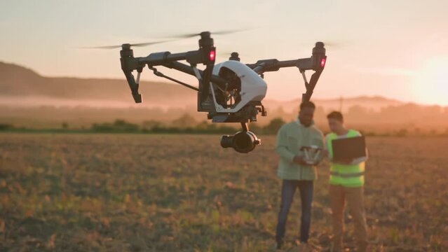 Agribusiness innovations. Flying drone in agricultural farm. Two multi-ethnic engineers control copter with tablet and laptop, checking soil and harvest.