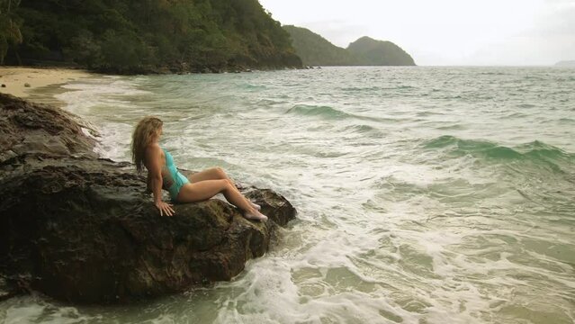 Woman chill relax resting on rock of sea reef stone, stormy cloudy ocean. Woman in swimsuit dress tunic. Concept rest tropical outdoor tourism