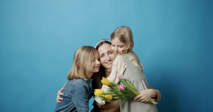 Happy Mom hugs daughters and kisses, with flowers, posing isolated on a blue background. Sisters with tulips flowers give on Mother's Day. Embracing love for 8 March. Happy Mother.