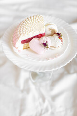 A white-pink piece of cake lies on a white plate on a light background. A pink heart-shaped cake lies on a white plate on a light background. Light macaroons on a white canvas. Delicious pastries