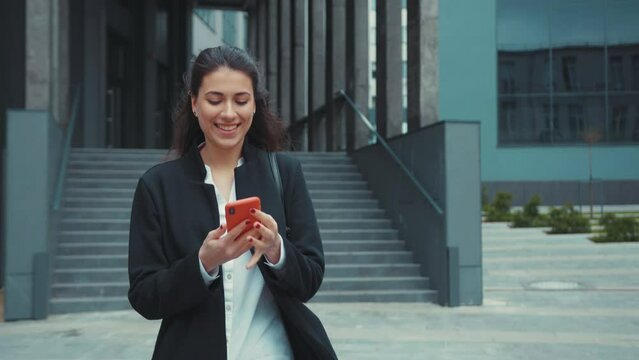 Happy attractive female executive leaving office building answering phone call. Cheerful caucasian young lady speaking mobile conversation on smartphone outdoors.
