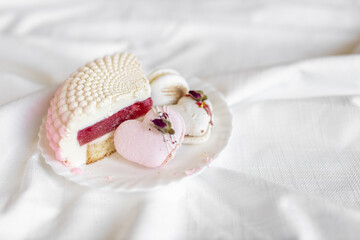 A white-pink piece of cake lies on a white plate on a light background. A pink heart-shaped cake lies on a white plate on a light background. Light macaroons on a white canvas. Delicious pastries