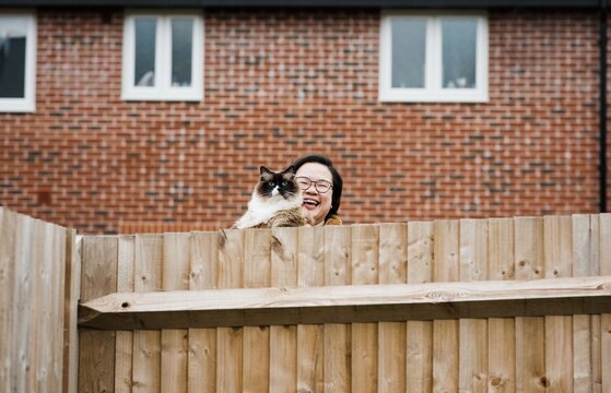 Woman With Her Cat Smiling Over Garden Fence Saying Hello To Neighbour