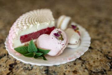 A white-pink piece of cake lies on a white plate on a light background. A pink heart-shaped cake lies on a white plate on a light background. Light macaroons on a white canvas. Delicious pastries