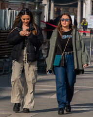 Two young women walking in the city; they are both wearing stylish outfits. One is wearing sunglasses, the other is looking at her phone.