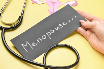 Woman holding paper with word MENOPAUSE, stethoscope and uterus on yellow background, closeup