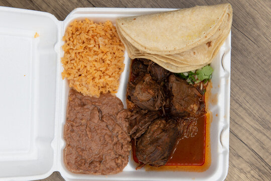 Overhead view of hearty take out order of birria stew meat served to make tacos with the included tortillas, rice and beans in the styrofoam box