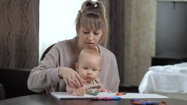 Young mother teaches cute baby son to draw with colored markers on paper sheet. Family adheres to early education sitting at table in bedroom at home. Mom, baby play together and draw at home in room