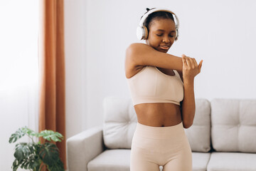 Fototapeta na wymiar Young black woman in headphones listens to music and does workout at home in the living room. Home training.
