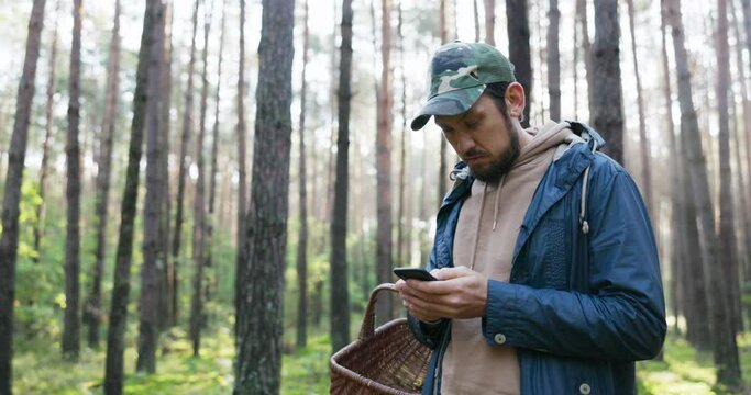 Young man mushroom picker walking in unknown forest looking for mushrooms stands in middle of trees with basket and phone, trying to get range, no signal, compass, navigation, lost, can't find way out