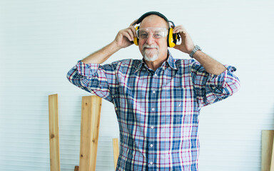 Senior Caucasian retired old happy male woodworker smiling, wearing check shirt, earmuff to protect loud noise for safety, create DIY wooden furniture for house decoration. Hobby, Retirement Concept. - obrazy, fototapety, plakaty