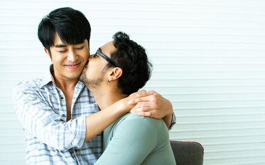 Portrait Gay LGBT sweet Asian couple wearing pajamas, smiling, hugging, kissing with happiness and love, sitting in living room at home with comfortable, posing in Lifestyle Concept.