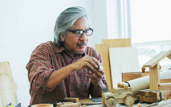 Senior Asian happy retired old male woodworker or carpenter smiling with happiness, wearing check shirt and eyeglasses, creating DIY wooden model for house decoration. Hobby, Retirement Concept.