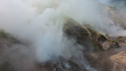 A geyser erupts on a mountainside. Splashes of boiling water are visible. Everything is shrouded in thick steam.  The river flows along a rocky bed. Kamchatka. Valley of Geysers. Poor visibility.