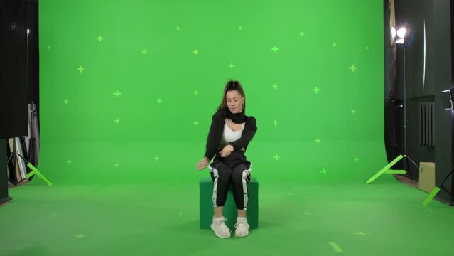 A young woman dancing on a green screen background. Attractive girl making content for social media. Female danser sits on box. Chroma key