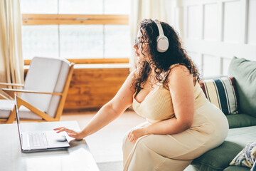 Curly haired overweight young woman wearing comfortable clothes in wireless headphones using laptop on sofa