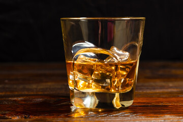 Half-poured glass of whiskey and ice on dark oak boards on black background. Close-up.