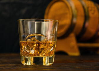 A half-filled glass of whiskey with ice cubes stands in front of an oak barrel.