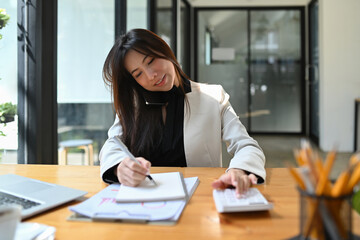 Busy businesswoman talking on mobile phone and using calculator at office desk.