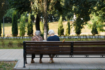 Back view of two old caucasian women girlfriends relaxing in park on summer day sitting on bench...