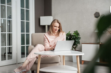 An attractive middle-aged woman in home clothes sits at home in a chair in front of a laptop. Freelancing and distance learning. Work in quarantine.