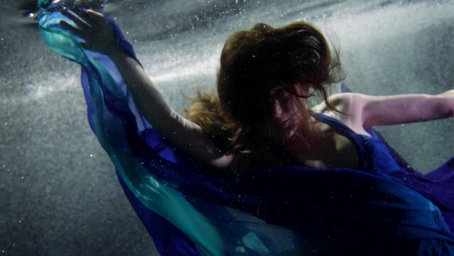 graceful lady in blue silk dress is swimming underwater, subaquatic shot in pool or aquarium