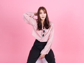 Portrait studio shot of Asian young beautiful hipster female teen model in casual street wears sneakers and sunglasses posing smiling look at camera sit on white square box stool on pink background