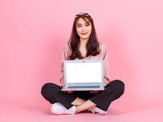 Naklejka premium Portrait studio shot of Asian young hipster female model in casual street wears sunglasses sit on floor holding pointing white blank empty screen laptop computer for advertising on pink background.