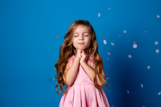 cute little girl with curls in a pink dress makes a wish and dreams standing with her eyes closed. studio background on a blue background. space for text. High quality photo