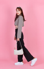 Portrait studio shot of Asian trendy fashionable female hipster teen model in casual crop top street wears jacket sunglasses sneakers holding handbag purse walking look at camera on pink background