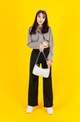 Portrait studio shot of Asian trendy fashionable female hipster teen model in casual crop top street wears jacket sunglasses sneakers holding handbag purse look at camera on yellow background