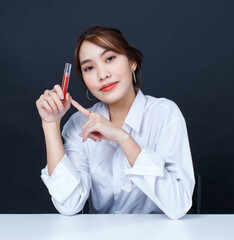 Portrait closeup studio shot of Asian young glamour trendy urban fashionable female model wearing makeup in casual white shirt sitting smiling look at camera holding red lipstick on black background