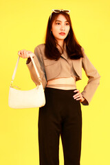 Portrait studio shot of Asian trendy fashionable female hipster teen model in casual crop top street wears jacket sunglasses sneakers holding handbag purse look at camera on yellow background