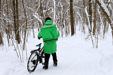 Delivery service courier with a thermos bag and bicycle by the snow in a park. Food delivery home in winter
